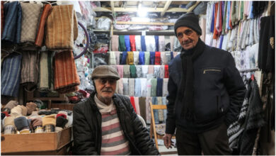 بائعان سوريان يجلسان خارج متجر لبيع الملابس على طول أحد أزقة السوق المغطى في مدينة حمص القديمة