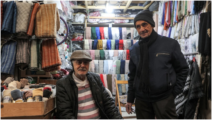 بائعان سوريان يجلسان خارج متجر لبيع الملابس على طول أحد أزقة السوق المغطى في مدينة حمص القديمة